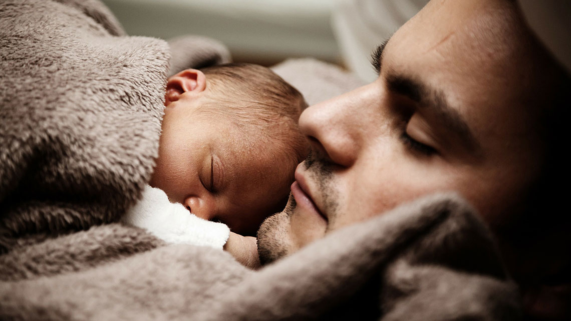 Baby schläft unter eine Decke gekuschelt auf dem Bauch des Vaters.