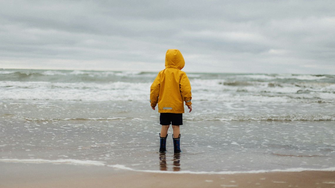 Kind mit gelber Regenjacke und Gummistiefeln steht am Strand, knöcheltief im Meer. | © unsplash
