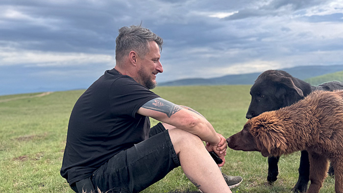 Gerd Schuster sitzt auf einer Wiese, zwei Hunde sitzen vor ihm. | © privat