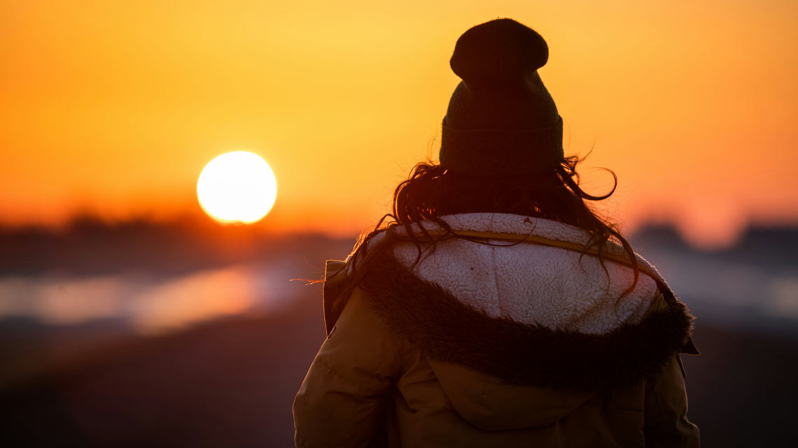 Warm angezogene Frau von hinten, die in der Ferne den Sonnenuntergang beobachtet. | © unsplash