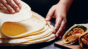 Wraps, die von zwei Händen gefaltet werden. Daneben zwei gefüllte Wraps auf Holzbrett. | © Massimo Pessina