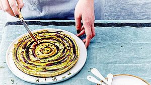 Eine Frau steht vor einem Tisch mit einer Gemüse-Tarte und schneidet diese mit einem Messer an.