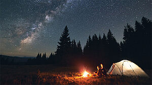 Zwei Personen am Lagerfeuer neben einem Zelt, Nachthimmel mit Sternen. | © anatoliy_gleb; Shutterstock.com