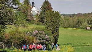 Pilgergruppe, die über einen Feldweg umgeben von Wäldern und Wiesen Richtung Kirche läuft. | © Belinda Gagern