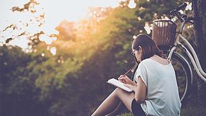 Frau sitzt im Sonnenuntergang neben ihrem Fahrrad im Gras, sie schreibt Tagebuch | © Teechai, shutterstock.com